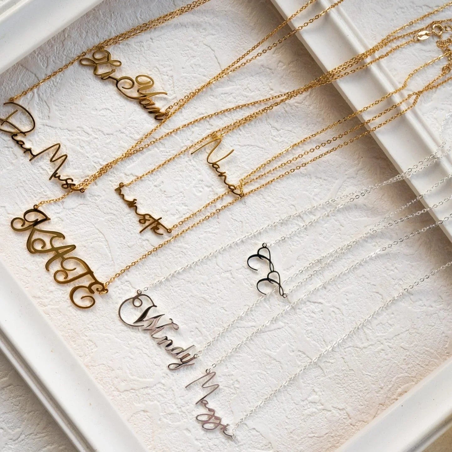 Personalized gold and silver name necklaces displayed on a white textured surface in a decorative frame