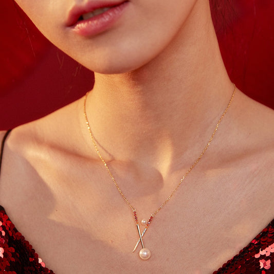 Close-up of a woman wearing a delicate gold necklace with two crossed pins adorned with red gemstones and pearls