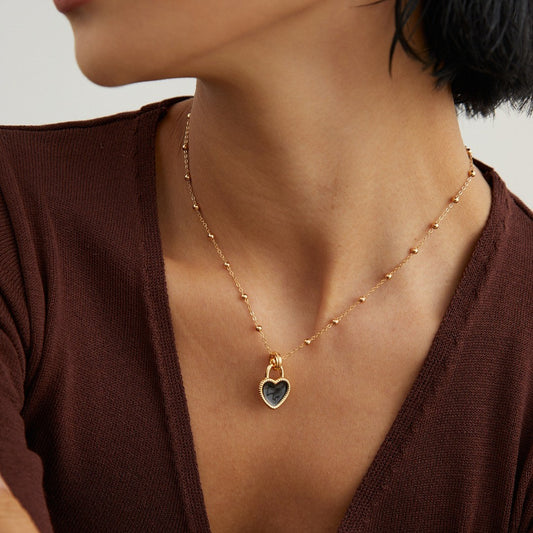 Close-up of a woman wearing a gold beaded chain necklace with a heart-shaped black pendant, styled with a brown top