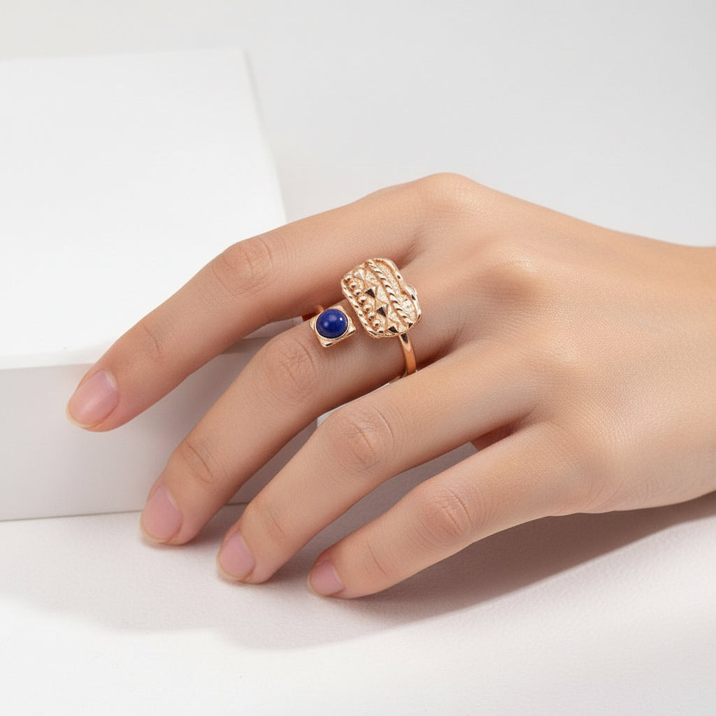 Hand wearing a gold ring with a textured rectangular design and a blue gemstone on a white background