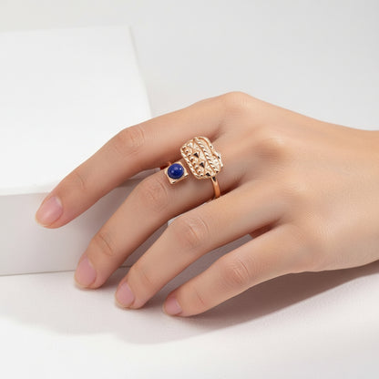 Hand wearing a gold ring with a textured rectangular design and a blue gemstone on a white background