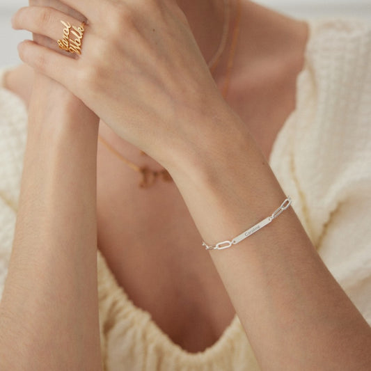 Close-up of a woman's wrist wearing a silver name bracelet engraved with Caroline and hands wearing gold rings