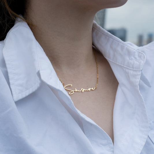 Close-up of woman wearing gold custom name necklace 'Singmore' with white collared shirt outdoors